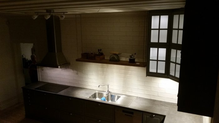 Mörkbild av kök under renovering med LED-spottar, trähylla och rostfri diskbänk.