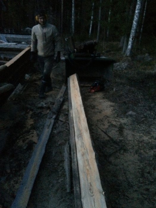 Person står bredvid stockar och en motorsåg i skymning, byggarbetsplats i skogsmiljö.