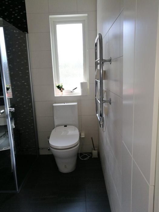 Toalett i ett renoverat badrum med vita och gråa kakelväggar, fönster och handdukstork.