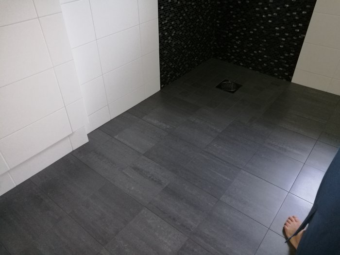Gråa klinkergolv och vita kaklade väggar i ett badrum med en svart mosaikduschhörna.