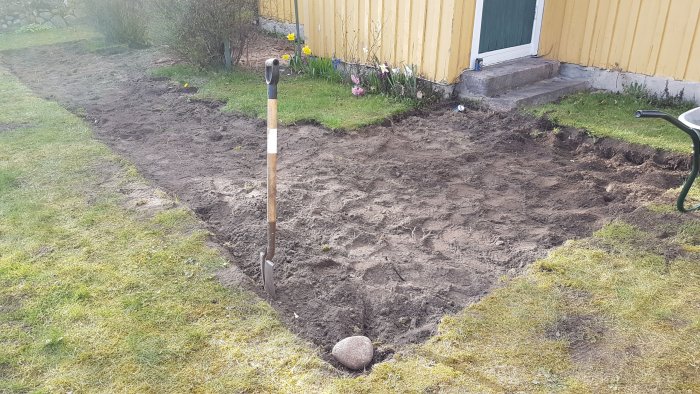 Påbörjad grävning av mark för altanbygge nära ett gult hus med en spade plantad i jorden.