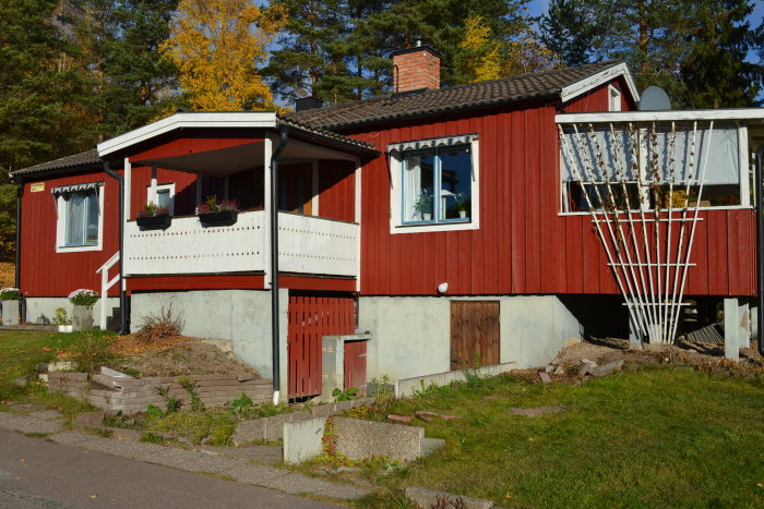 Rödmålat hus med vit knut och balkong inför trädgårdsrenovering, omgiven av träd och gräs, med rensad entré.