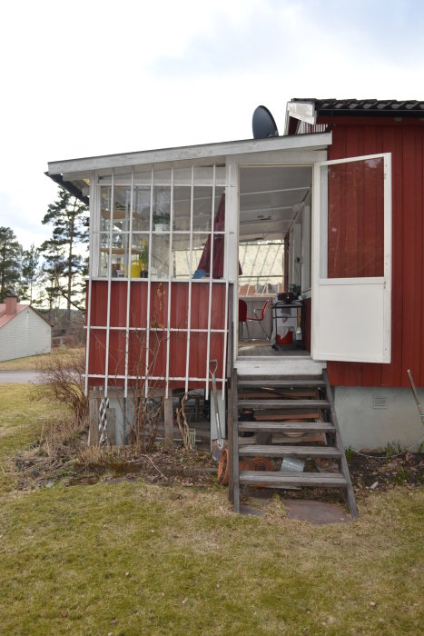 Ingång till ett hus med en veranda omgiven av glaspaneler och röda väggar, rensad från växtlighet.