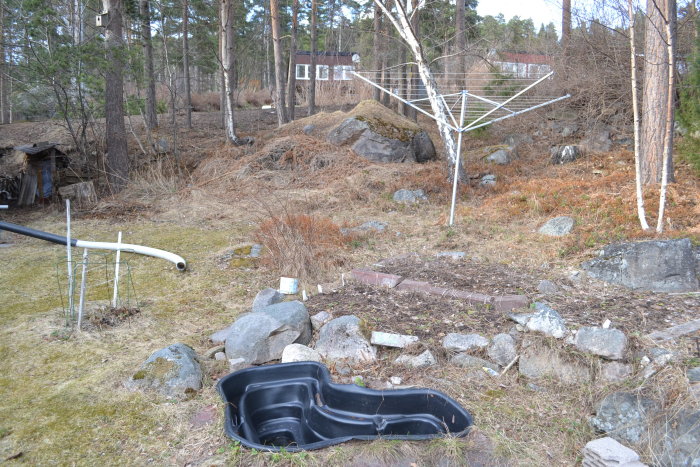 Uppträdgård med vissna växter, en torkställning och en tom damm, med skog och stenar i bakgrunden.