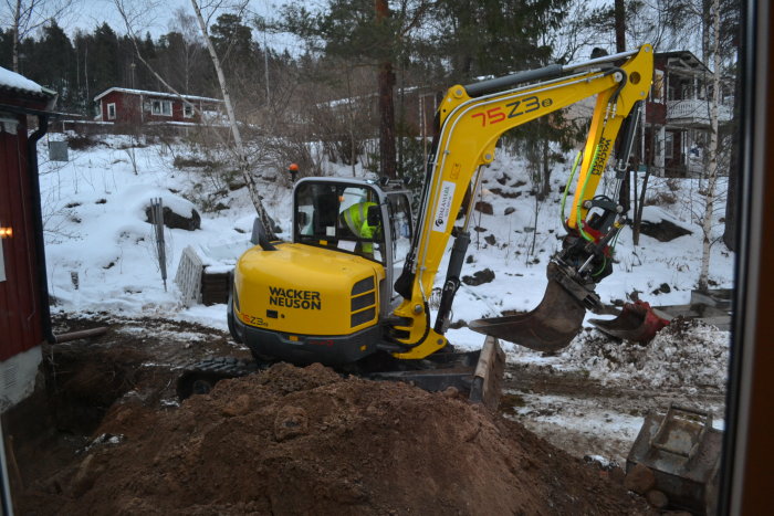 Gul grävmaskin, Wacker Neuson, gräver i en trädgård med snötäckta markytor och hus i bakgrunden.