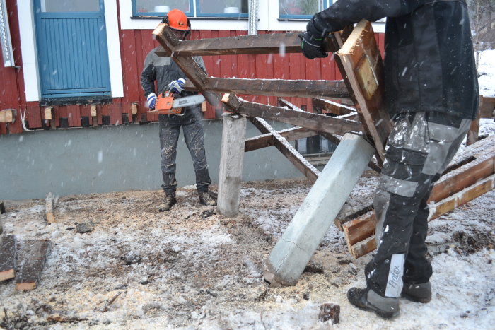 Två personer demonterar ett träuterum i snöfall, en sågar i en träbjälke medan den andra håller en stående pelare.