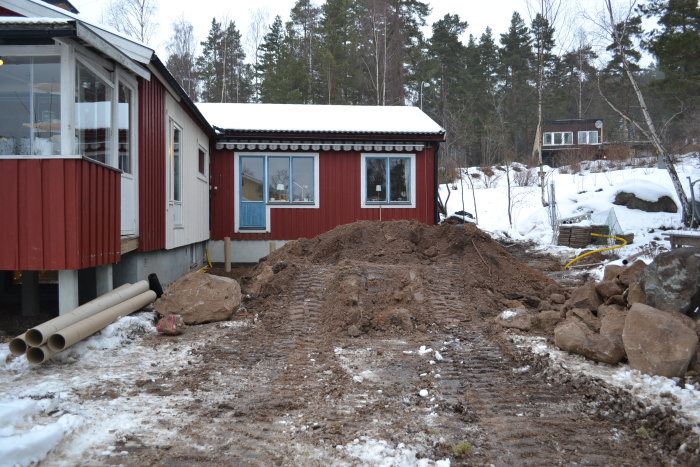 Hus med uppgrävd mark för dränering, byggmaterial och snö syns, med skog i bakgrunden.