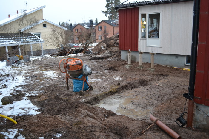 En grävd och lerig trädgård med en betongblandare framför ett hus med stöttor undergrund.