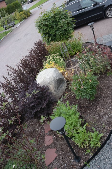 En omsorgsfullt planterad perennrabatt med blandade buskar, en stor sten och dekorativa trädgårdslampor.