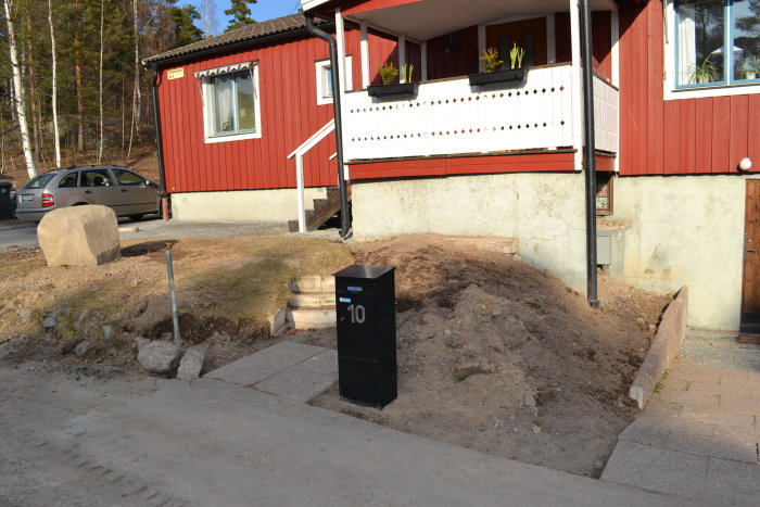 Provisorisk trappa av trä och sten framför en röd husfasad, med en nyplöjd rabatt och en svart brevlåda vid vägen.