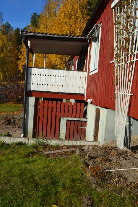 Röd trähusfasad med vit balkong och en improviserad trätrappa vid en nyanlagd rabatt under höstfärgade träd.