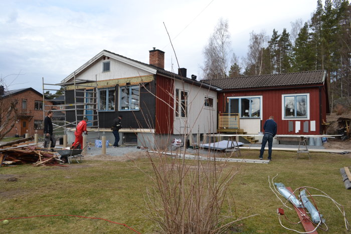 Renovering av uterum vid ett rött hus med byggarbetare, ställningar och byggmaterial på tomten.