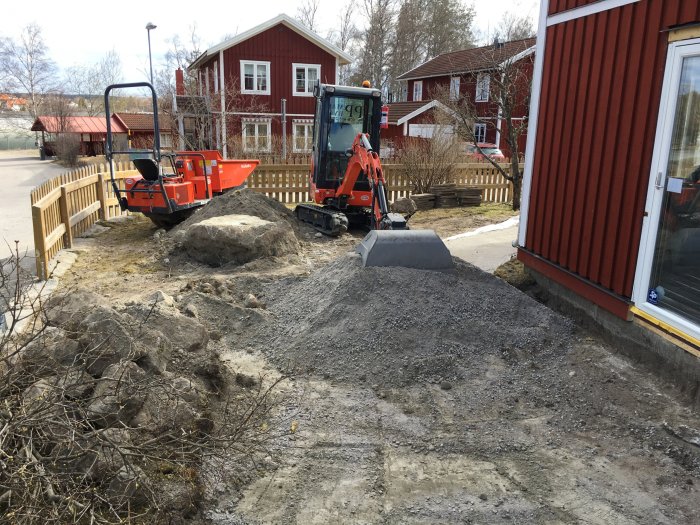 Utomhusarbeten med en grävmaskin och en dumpervagn bredvid en stor hög med grus på en byggarbetsplats.