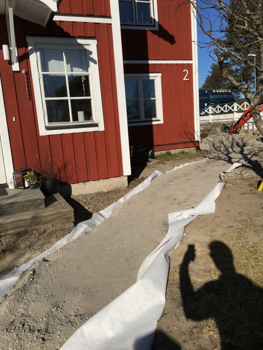 Rödfärgat hus med nummer 2 och nygjuten gångväg kantad av vit fiberduk, skugga av en person syns.