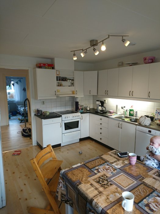 Kök före renovering med vita skåp, spis, bordsduk och lekande barn på golvet.