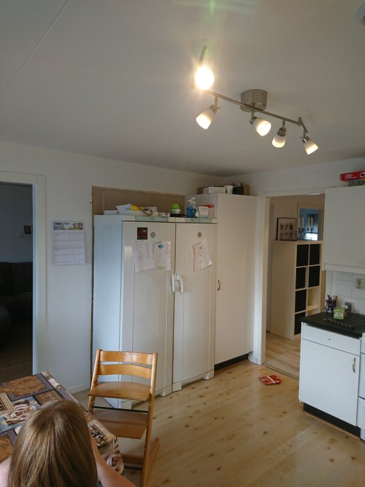 Ett kök före renovering med vitvaror och en barnstol framför kylskåpet.