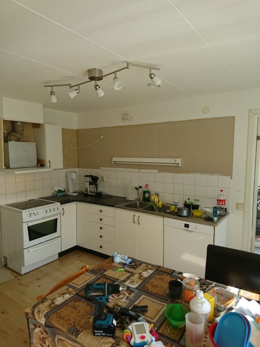 Kök under renovering med borttagna överskåp och synliga vägginstallationer samt verktyg och leksaker på matbordet.
