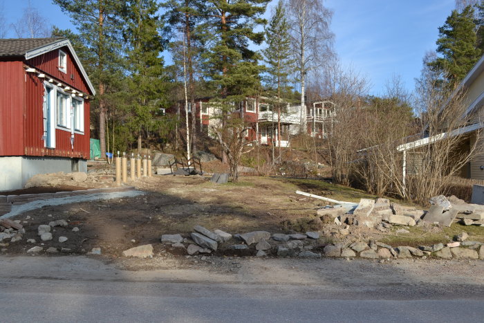 En oordnad tomt med stenar och byggmaterial, en påbörjad stenmur längs gatan, och planterade stolpar i bakgrunden.