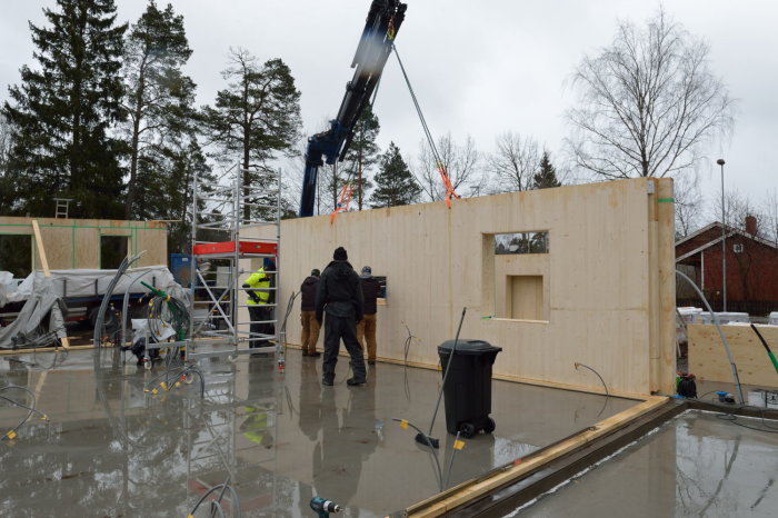 Första prefabricerade träväggen monteras på byggarbetsplats med kranbil och arbetslag.