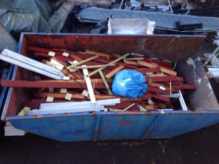 En container full med byggavfall, inklusive röda läkter och vit panel.