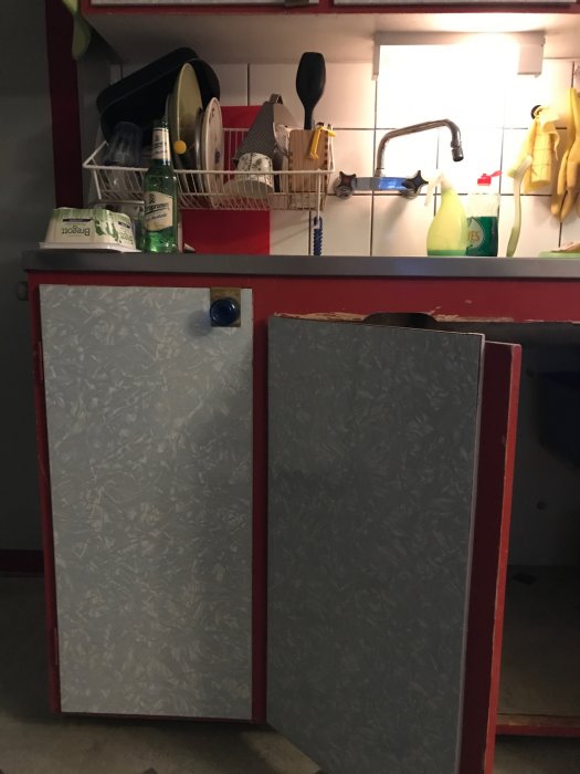 Ålderstiget kök med diskbänk och upptorkad disk, ett kallvattenrör synligt under diskbänken till höger.