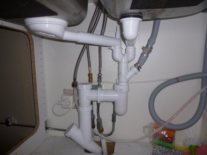 Rörinstallation under köksvasken med synliga vattrör, avlopp och sladdar.