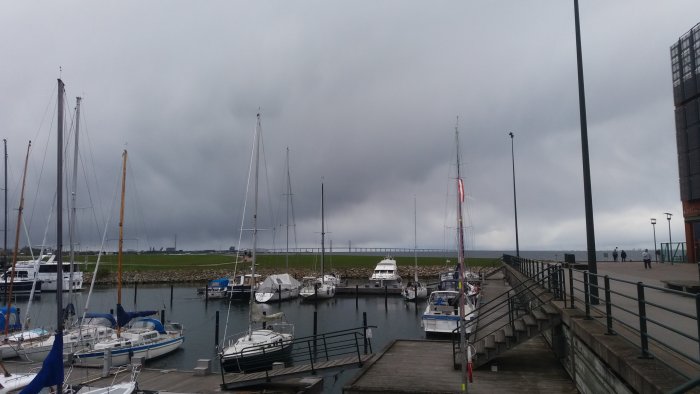 Mörka moln hänger över en småbåtshamn med segelbåtar, vy mot en gräsmark och byggnad i bakgrunden.