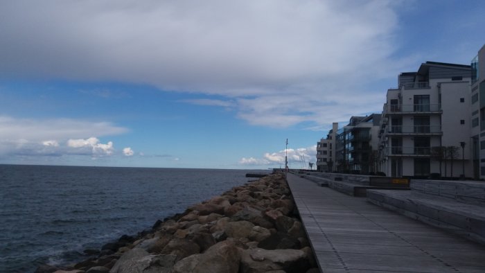 Solig vy över havet med moderna vitfärgade byggnader längs en kaj och stenmur.