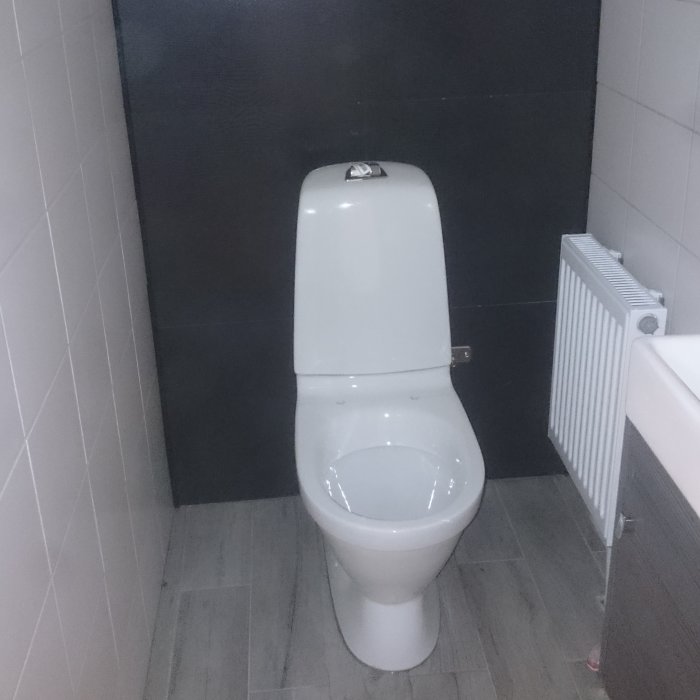 Renoverat badrum med ny monterad vit toalett, vita och mörka kakelväggar samt en vit radiator.