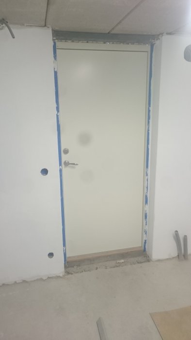 Nyinstallerad vit dörr med dörrhandtag och låscylinder, omgivet av ofärdig vägg och skyddsplast.