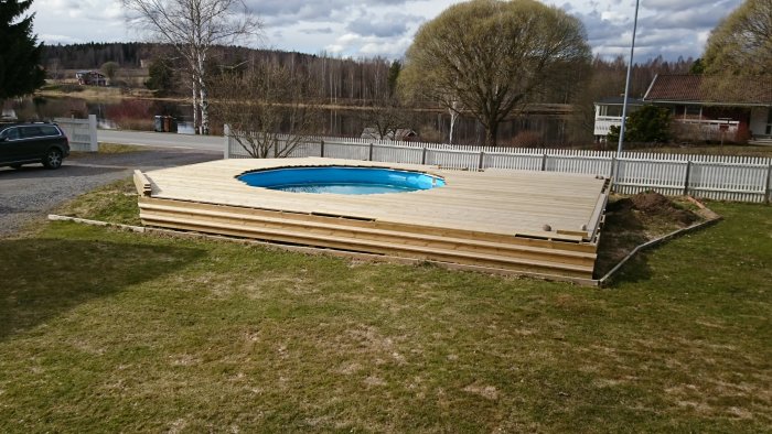 En halvfärdig träterrass runt en inbyggd blå pool med vintergräs och staket i bakgrunden.