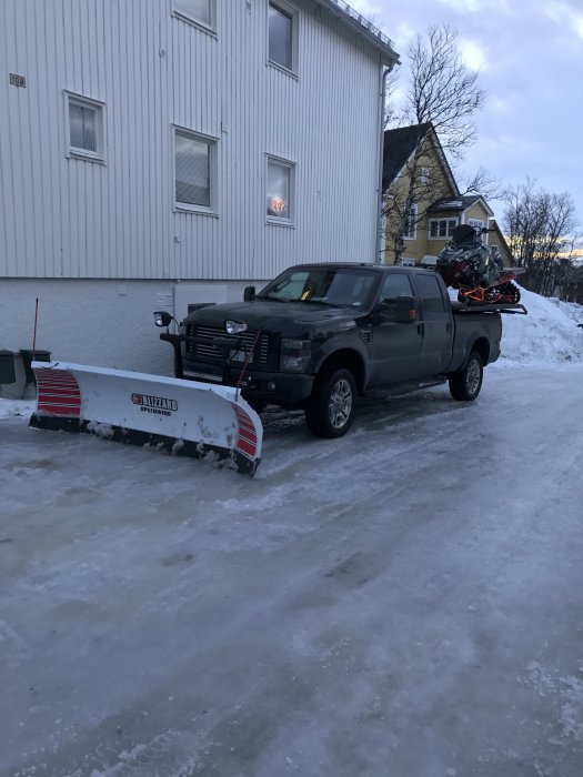 Pickup med snöplog fram och snöskoter på flaket parkerad på isig väg vid hus