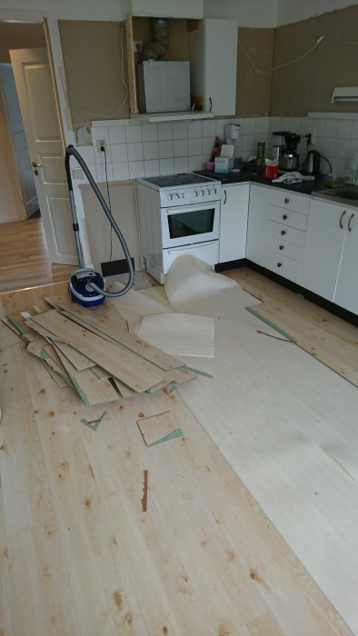Kök under renovering med borttagna laminatgolv, utsatt gipsskiva och dammsugare på plats.