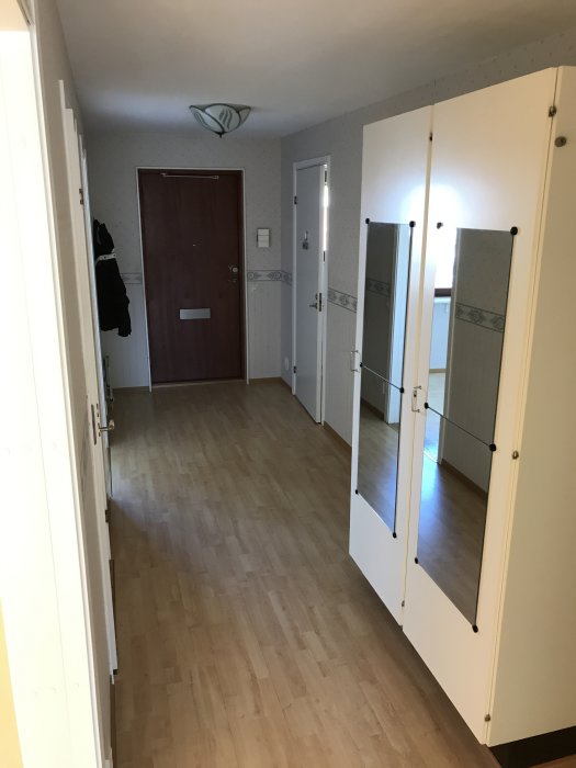 Hall före renovering med vita garderober längs väggen och en brun dörr i bakgrunden.