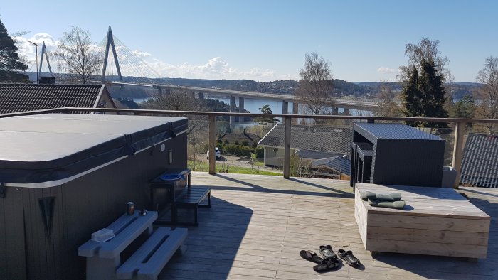 Träaltan med sittmöbler och bubbelbad, glasräcke, utsikt över en bro och ett landskap med vatten och himmel.