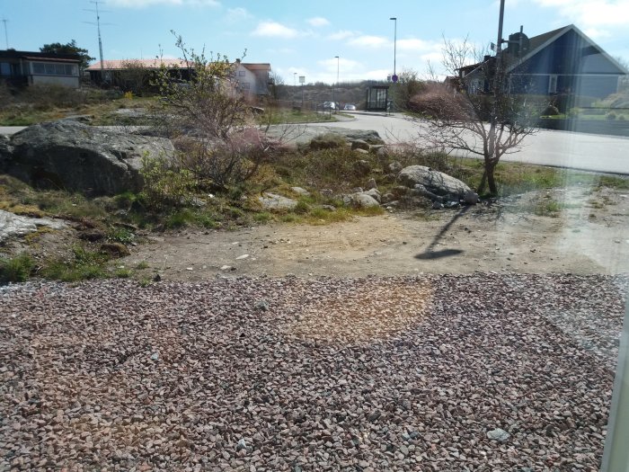 Tomt med grus och lite växtlighet intill trafikerad väg och hus i bakgrunden.