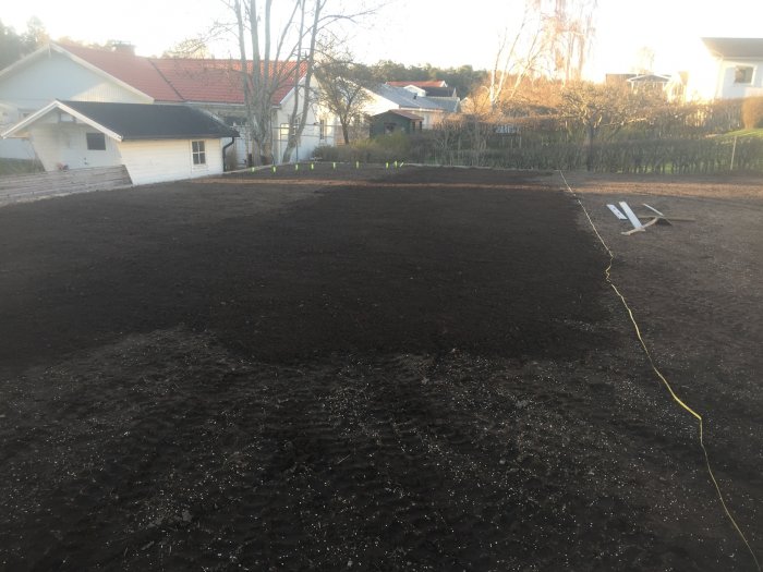 Nyplanterad trädgård med finjord och stenrad, verktyg på marken, hus och träd i bakgrunden.