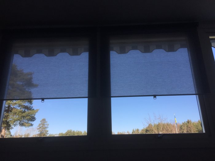 Två nedfällda rullgardiner i ett vardagsrum med utsikt mot träd och klarblå himmel.