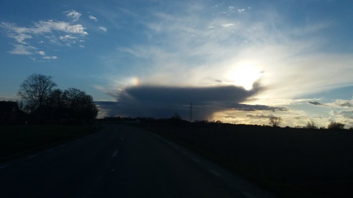 Himmel och moln med en liten regnbåge synlig bredvid en stor molnmassa, sett från en landsväg vid skymningen.