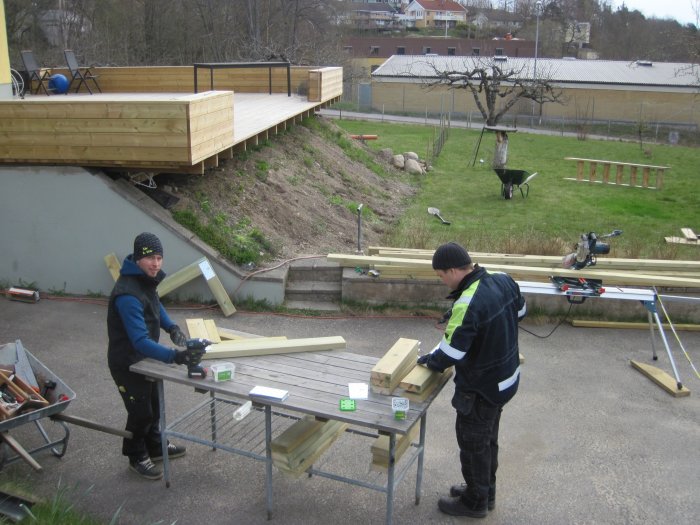 Två män arbetar med träkonstruktioner, färdig altan och stödstrukturer i bakgrunden.