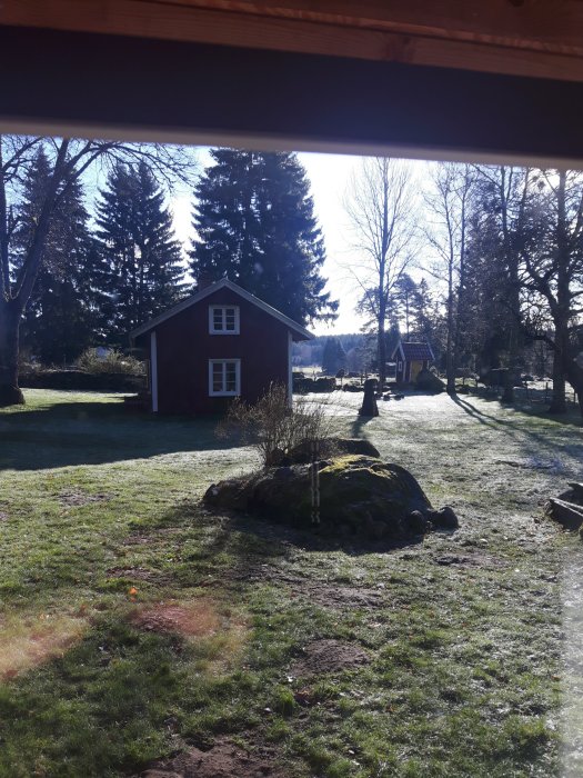 Röd stuga med vita knutar omgiven av träd och gräsmatta, delvis i skugga, fotograferad från halvtäckt veranda.