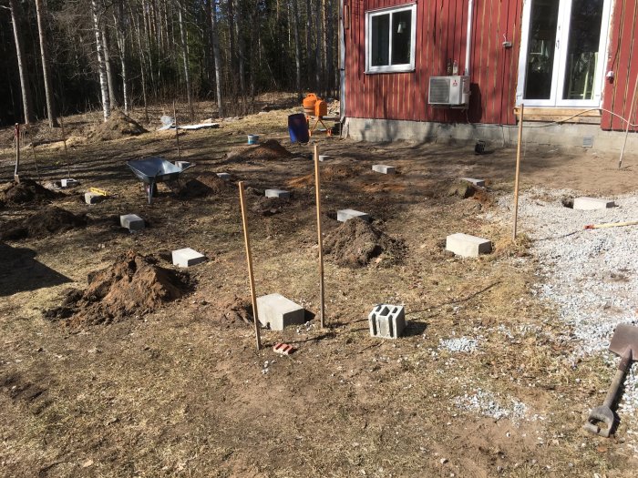 Byggarbetsplats utomhus med nyligen installerade betongplintar och trästolpar i marken vid ett hus med röd fasad.