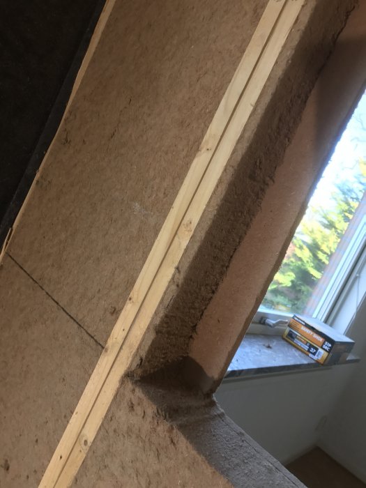 Vinkelvy av inre hörn i ett rum under renovering med osb-skivor och träreglar på en vägg och synligt mörkt isoleringsmaterial.