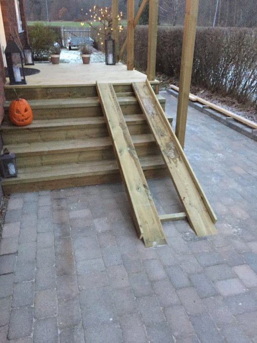 Trätrappa och barnvagnsramp av trä på en uteplats med stolpar i bakgrunden och snöfläckar.