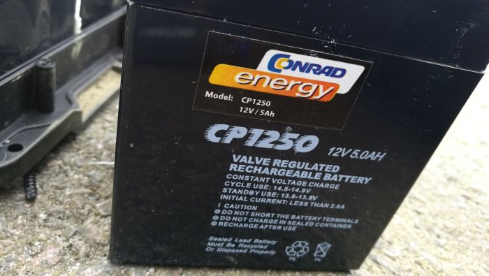 Conrad energy 12V 5Ah blybatteri modell CP1250 med specifikationsetikett mot grusig bakgrund.