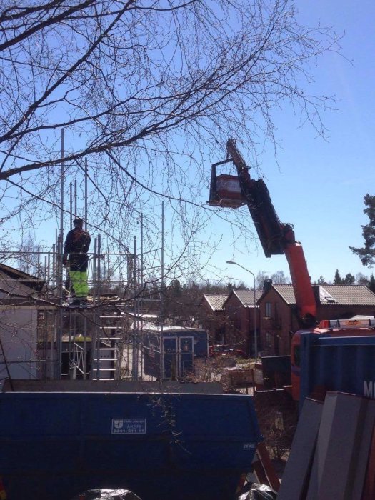 Arbetsplats med lyftkran och arbetare på byggnadsställning vid takläggningsprojekt, med material på marken.