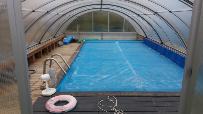 Pool täckt med bubbelfolie inomhus under ett stort, halvöppet växthusliknande pooltak.