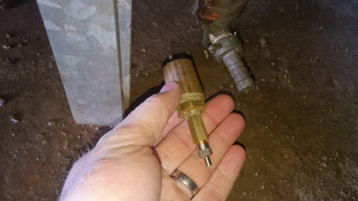Hand håller en nippel för VVS-användning nära rostiga rörledningar på ett betonggolv.