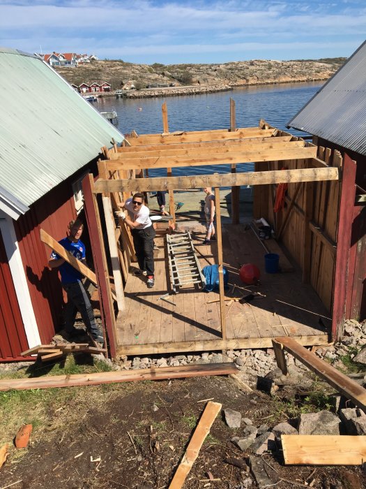 Personer som river en brygga vid en sjöbod med havet och himmel i bakgrunden.