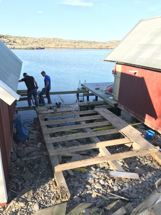 Två personer rivit en träbrygga vid havet med delar av bryggan och bråte på marken.